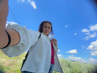 A young woman is taking a selfie outdoors on a sunny day, with a bright blue sky and scattered white clouds above. She is wearing a white open shirt over a pink top and blue jeans. She has sunglasses perched on her head. The background shows a grassy hillside with some bushes and scattered power lines. The overall scene appears to be in a rural or suburban area.