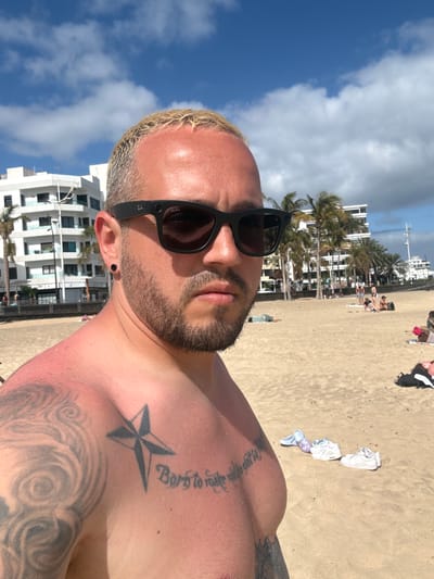 A selfie of a man at Playa del Reducto, Arrecife, Lanzarote, Spain. The man has a blonde buzzcut, beard, tattoos on his shoulder and chest, and is wearing sunglasses. Behind him is the beach with other people relaxing, some buildings, palm trees, and the sky with clouds.