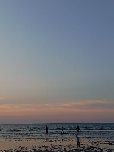 The image displays a coastal scene in the city of Tritunggal, Indonesia, during either dawn or dusk. The sky exhibits a gradient from a light blue-grey at the top to soft orange-pink and purple tones near the horizon, where subtle, elongated cloud formations are visible. The calm, dark blue-grey sea meets the sky at a clear horizontal line. The foreground is composed of a wide expanse of dark, wet sand, heavily reflecting the sky's colors and the figures, indicating a low tide or intertidal zone with discernible patterns from receding water. Three human figures are present, partially silhouetted, walking or standing in the shallow water along the shoreline. The leftmost figure wears dark clothing, with a possible reddish upper garment. The central figure walks from left to right, wearing light-colored trousers. The rightmost figure stands, appearing to hold a smaller individual or object, with a clear reflection visible on the wet sand.
