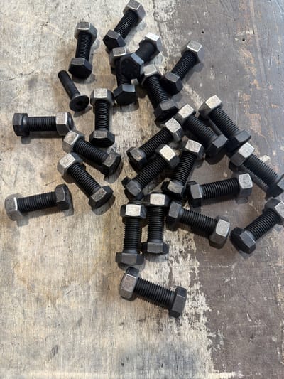 The image displays a collection of hexagonal head bolts and one Allen head bolt scattered across a weathered, textured wooden surface. The bolts are primarily black with silver-colored hexagonal nuts on many of them. The wood surface appears to be a workbench or floor, with visible grain, scratches, and what may be oil stains or a patina of age. The lighting suggests it is daytime, with shadows cast by the bolts indicating an overhead light source. The scene is a close-up, top-down view of these hardware items. There are no people present. The arrangement of the bolts appears random, as if they have been dropped or are waiting to be used. The overall impression is one of a workshop or maintenance area. There is no discernible text visible in the image.