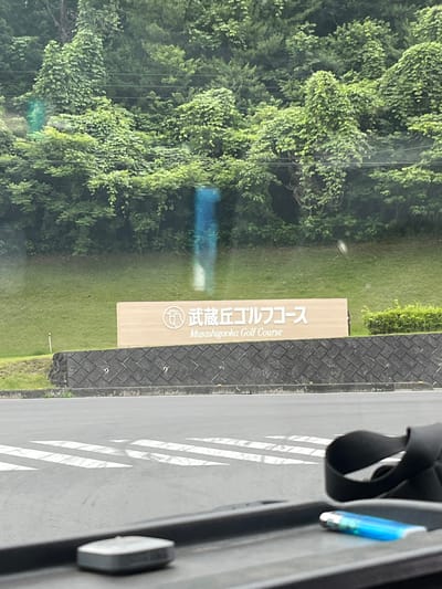 The image shows a sign for "Musashigaoka Golf Course" in Hanno, Japan. The sign is light brown with white lettering and a circular logo. Behind the sign is a grassy hill covered in lush green trees. In the foreground, a road with white crosswalk markings is visible. A black strap and a blue lighter are resting on a dark surface in the lower right corner of the image, suggesting it was taken from inside a vehicle.