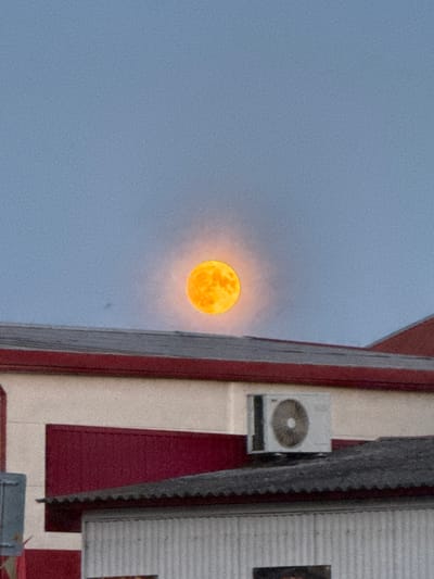 A full, orange moon is visible in a pale blue sky, surrounded by a soft halo of light. It is positioned above the roofline of a building, casting a warm glow. The building has a white facade with red trim and a dark, corrugated roof. An air conditioning unit is mounted on the exterior wall of the building. The lower portion of the image shows another part of the building with a light-colored, ribbed exterior. The image appears to be taken in an urban or suburban setting, likely in the city of Cherven, Belarus, given the context.