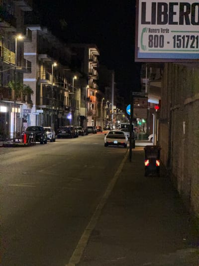The image depicts a street scene at night in Novara, Italy. Buildings line the left side of the street, illuminated by streetlights and interior lights. Several cars are parked along the curb on the left and are also present in the street, with one white car prominently featured in the middle of the road. On the right side of the image, a brick wall forms the boundary of a building. A trash receptacle with reflective markings is visible on the sidewalk to the right. A large billboard with the word "LIBERO" and a phone number is mounted on a building facade on the right side of the street. The street itself is paved, with visible lane markings. The overall impression is of a quiet, urban night.