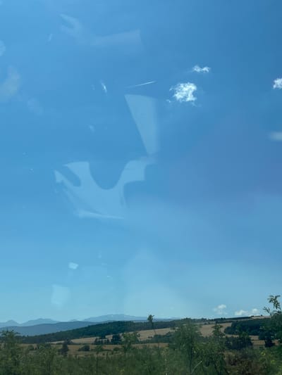 The image shows a bright blue sky with scattered white clouds. There are reflections on the surface of the sky, suggesting it might be viewed through a window. In the distance, there are rolling hills and mountains, covered with a mix of trees and fields. The terrain appears to be rural and somewhat arid. The colors are muted, with shades of green, brown, and blue dominating the scene. There are no people or animals visible in the image. The overall impression is one of a serene, natural landscape on a clear day. The exact location in Vakarel, Bulgaria, cannot be determined from the image alone, but the scenery is consistent with a rural setting in that region.