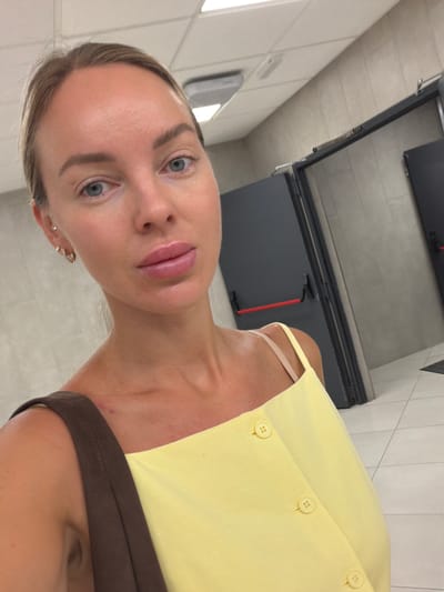 In a brightly lit restroom, a young woman with blonde hair pulled back takes a selfie. She is wearing a yellow sundress with delicate straps and three pearl buttons down the front. A brown strap from a purse rests on her shoulder. Her face is turned towards the camera, with blue eyes and full lips. She has small hoop earrings in her ears. The background shows tiled walls and two dark gray doors with red push bars, typical of public restrooms. The floor is tiled in a light color. The setting appears to be a modern public facility, possibly in a commercial area or transportation hub within Villajoyosa, Spain, given the detail provided about the city.