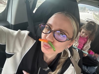 The image displays two females within a moving vehicle, identified as being in Menteşe, Turkey. In the foreground, a female with blonde hair and dark-framed oval eyeglasses is visible, facing forward. An orange and green gummy worm is positioned horizontally between her upper lip and nose. She wears a black jacket featuring beige and white panels. A light brown, zippered shoulder bag is partially visible on her right side. In the background, seated in the rear passenger seat, a young female child with dark hair in a ponytail is observed. She is looking down at an open book or tablet held in her lap, with a doll partially visible beneath it. The external environment, seen through the vehicle's rear window, shows a multi-lane road with distant vehicles, flanked by vegetated hills.
