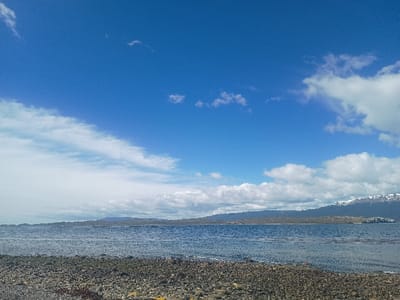 A bright blue sky with scattered white clouds stretches over a serene seascape. A rocky shoreline in the foreground gives way to calm waters. In the distance, landmasses, possibly islands or coastal mountains, are visible, with some peaks displaying patches of snow. The light suggests a clear, sunny day. The overall impression is one of a peaceful and remote natural environment.