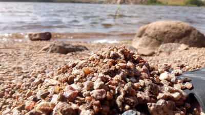 The image depicts a close-up of a pile of small, multi-colored pebbles on a shore, with a body of water and a distant shoreline in the background. The pebbles are the primary focus, appearing rough and varied in texture and color, ranging from light tan to reddish-brown and grey. Behind the pebbles, the water is visible, with gentle ripples reflecting sunlight. Several larger stones are scattered on the shore and in the shallow water. In the distance, the far shore appears to be lined with vegetation. A fishing rod, with a green and yellow tip, is partially visible in the water, suggesting the activity of fishing. The overall impression is a natural, outdoor scene, likely at a lake or river. The image does not contain any identifiable landmarks or features that would definitively place it within the city of Buzke, Ukraine.