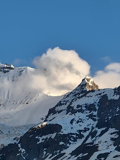 The image depicts a majestic mountain range under a clear blue sky, with scattered white clouds. The peaks are covered in snow, with dark rocky outcrops visible through the snow cover. A prominent peak in the foreground is bathed in sunlight, highlighting its sharp, triangular shape. Clouds are interacting with the mountain terrain, obscuring parts of the landscape and creating dramatic lighting effects. The scene suggests a high-altitude, rugged environment. The location is Kead, India, known for its mountainous terrain. There are no people or specific objects visible that would indicate human activity or a specific event. The environment is natural and grand.