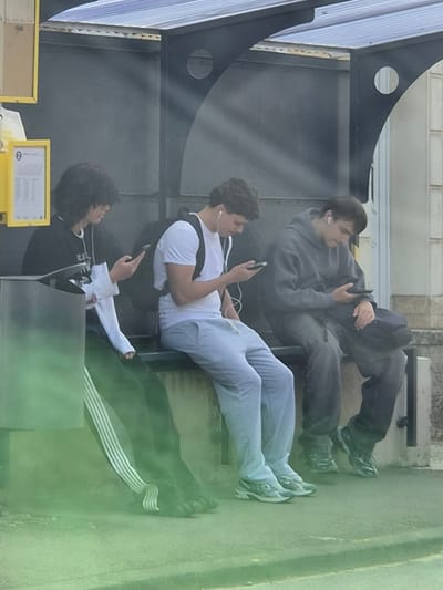 Three individuals are seated side-by-side on a bench, likely at a bus stop, in Sanem, Luxembourg. The person on the left, with dark, shoulder-length hair, wears a black long-sleeved shirt with a white graphic and green pants with white stripes down the side. They are looking at their phone, holding it in their left hand. A white sleeve from a garment underneath is visible.
The middle individual has short dark hair and wears a white t-shirt and light blue sweatpants. They are looking down at their phone, which they hold in their right hand. A backpack is visible behind them.
The person on the right, with short brown hair, is wearing a grey hooded sweatshirt and dark pants. They are looking at their phone, which they hold in their right hand, and have white earbuds in. A dark bag rests on their lap.
The background appears to be a bus stop shelter with a metal roof and dark grey paneling. A yellow sign with a schedule is mounted on the wall to the left, and a circular detail is visible on the back panel of the shelter. The ground in front of the bench is a light grey surface, with a yellow line marking an edge, and is covered by a noticeable green haze, possibly due to a filter or lighting condition.