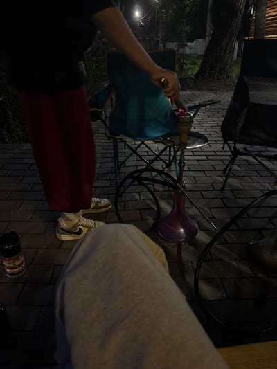 An overhead shot captures a person from the waist down, with their left leg extended and right leg bent. They are wearing a dark t-shirt and a dark red skirt, white socks, and white sneakers with black accents. The person's hand, holding a metal utensil, is positioned over a hookah. The hookah features a purple base and a metallic stem, with a charcoal-filled bowl at the top. A teal folding chair is partially visible to the left of the hookah, and another dark chair is to the right. The scene is set on a paved patio made of dark grey bricks. In the background, a large tree with rough bark stands prominently, its branches extending upwards. The lighting suggests it is nighttime, with some ambient light illuminating the scene. A black thermal mug with colorful bands is placed on the ground to the left of the frame. The overall atmosphere is relaxed and informal. The location is Doma 1096 Km, Russia.
