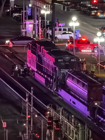 In Hollywood, California, a train, possibly a freight locomotive numbered 801, is stopped on railroad tracks at night. The train is illuminated by purple and pink lights, creating a dramatic visual effect. Several people are present near the train, including individuals in uniform who appear to be emergency responders. A red fire rescue vehicle with flashing lights is also visible on a nearby street. The scene suggests an ongoing incident or a planned event that has drawn emergency services. The surrounding area is dark, with streetlights and vehicle lights providing illumination.