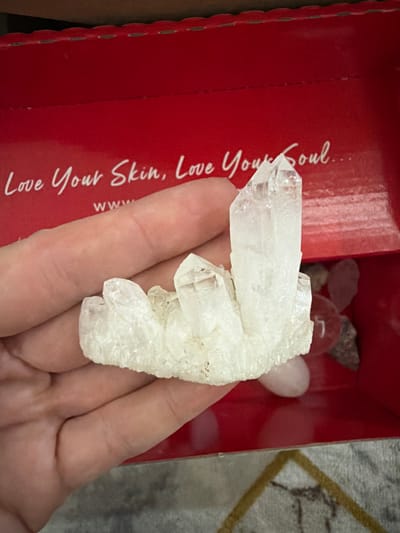 A close-up, indoor, eye-level shot shows a person's hand holding a cluster of clear quartz crystals. The crystals are milky white and translucent, with sharp, geometric facets. They are resting on a bright red, glossy box with white text that reads "Love Your Skin, Love Your Soul..." and "WWW". The background of the box is blurred, showing more crystals and possibly small stones. The lighting is warm and natural. The image does not provide any information about the location in İzmir, Turkey.