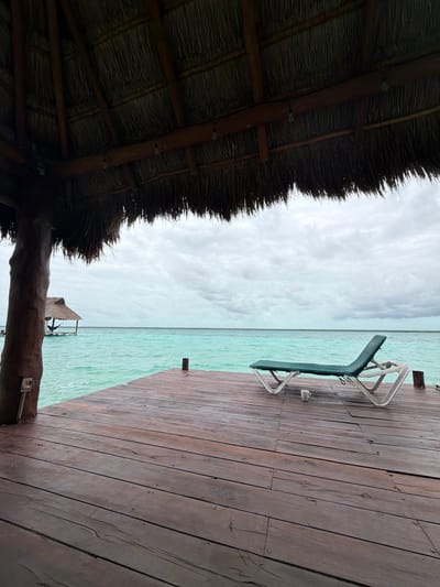 The image depicts a serene tropical scene from beneath a thatched roof overhang, looking out towards a turquoise body of water under a cloudy sky. In the foreground, a rustic wooden dock extends into the water. On the dock, a green lounge chair with a white frame is positioned facing the water, with a white mug resting on the wooden planks in front of it. To the left, a thick wooden pillar supports the overhang, and an electrical outlet is visible on its side. In the distance, a small thatched-roof structure sits on stilts in the water, with a hammock visible underneath its roof. The water is calm with gentle ripples, and the horizon is a clear line between the water and the sky. The overall impression is one of peaceful, isolated relaxation. The scene is consistent with locations found in tourist areas around Bacalar, Quintana Roo, Mexico, which features a prominent lagoon known for its vibrant turquoise waters.