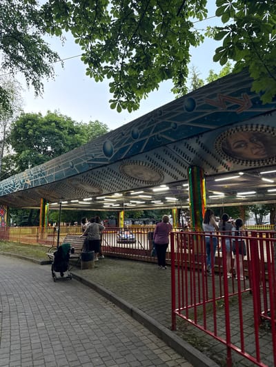 The image captures a scene at an amusement park, likely in Karaman 1-Yy, Russia, as indicated by the location context. The perspective is tilted, giving a dynamic view of a ride and its surroundings.
The main subject is a large, circular amusement ride with a decorative exterior featuring images of faces, celestial patterns, and illuminated lights. Several people are visible, either waiting in line behind a red metal fence or seated on the ride. Their attire suggests a casual outdoor setting. A woman in a purple top is standing near the fence, and another person, possibly a child, is seated on a bench with a stroller nearby.
The setting includes a paved walkway with a grey stone border, and lush green trees frame the top and left side of the image, suggesting it is daytime and likely a pleasant weather day. The sky is visible in the upper portion, appearing bright and clear, with a few birds flying in the distance.
The activity taking place is people queuing for or riding an amusement park attraction. The overall mood appears relaxed and typical of a recreational outing at an amusement park. There is no discernible text visible in the image.