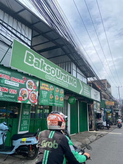 The image shows a street scene in Bojongsoang, Indonesia. A motorcyclist wearing a green helmet and black jacket with "Shopee Food" and "Grab" logos stands in front of a storefront with a large green sign that reads "Bakso Urat MG Outlet Bojongsoang" in white letters. There is another sign below that says "Special Promo Bakso Urat MG" in a green box. There are various other signs and objects on the storefront, including "Open 10:00-21:00" and "Tempat Makan Di Lantai 2 Halal." There are two other motorcycles parked in the street. The street has an asphalt surface, and there are many electrical wires overhead. The sky is partly cloudy. The location is a small town in the Bandung Regency, West Java, Indonesia.