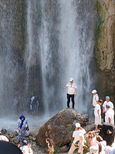 The image shows a waterfall in Arstanbap, Kyrgyzstan, with several people gathered around it. The waterfall cascades down a rocky cliff face, creating mist and spray. In the foreground, a woman in a blue and white patterned dress stands near the water's edge, holding a handbag. To her right, a young girl in a yellow and white patterned outfit looks towards the camera. In the mid-ground, a man in a white t-shirt and black pants stands on a large rock, gesturing towards the waterfall. Other individuals, some wearing white t-shirts and caps, are visible near the waterfall, observing the scene or interacting with each other. The environment is lush with greenery, and the rocks around the waterfall are wet and mossy.