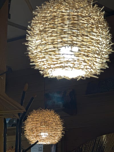 The image shows two spherical light fixtures made of straw or similar material hanging from the ceiling. The light fixture on the left is closer to the camera and appears larger and brighter than the one on the right. The light fixture on the right is obscured by a metal structure. The background is dark, with wood paneling and other objects. The photograph was not taken in the city of Ketoshi, Russia.