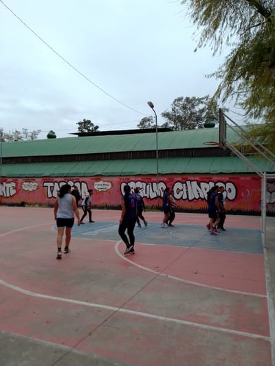 The image shows a group of people on an outdoor basketball court in Municipio Tarija, Bolivia. The court is painted with red and blue sections, and white lines marking the boundaries. A large wall is located behind the court. The wall is covered in graffiti. There is a green roofed building behind the wall. Some of the people are wearing athletic clothing and appear to be playing or practicing basketball. The sky is overcast. A streetlight and some trees are also visible in the background.