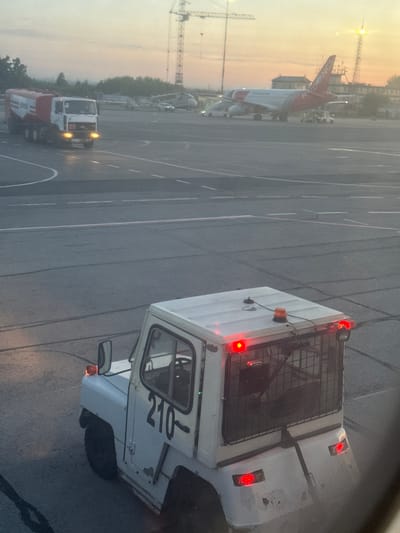 The image depicts a view from inside a window overlooking an airport tarmac. A white airport tug with the number "210" prominently displayed is in the foreground. Behind it, an aircraft with the word "REDWINGS" visible on its tail is present. A fuel truck is situated further in the background, and construction cranes can be seen on the horizon. The time of day appears to be dusk or dawn due to the lighting conditions. Given the mention of Kitsjanovo, Russia, it's likely this is Kitsjanovo Airport.