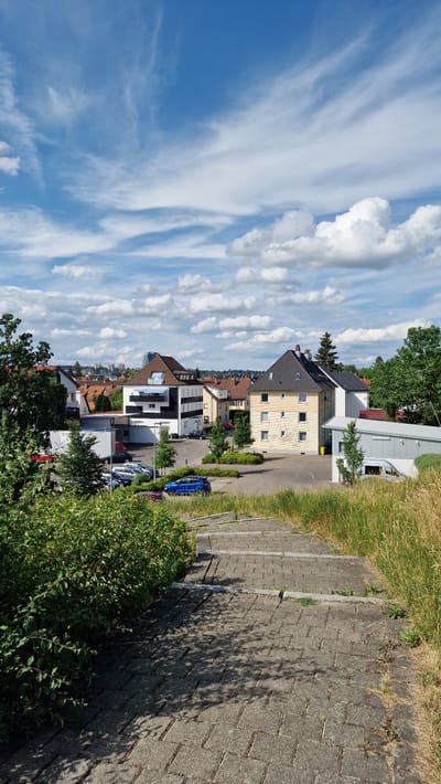 This is a landscape-oriented, rotated photograph of a residential street in Villingen-Schwenningen, Germany. The scene is bathed in bright sunlight, indicating it is daytime. The sky is a vibrant blue, filled with a mix of fluffy white cumulus clouds and wispy cirrus clouds, suggesting fair weather with some atmospheric movement.
The street is lined with multi-story houses, exhibiting a variety of architectural styles and colors. Some buildings appear older with tiled roofs and traditional windows, while others are more modern, featuring clean lines and large glass elements. A few of the houses have solar panels visible on their roofs, hinting at a focus on renewable energy. Greenery is abundant, with mature trees and lush bushes bordering the street and surrounding the houses.
A paved driveway runs along the right side of the image, with parked cars visible in the distance. One car, a bright blue hatchback, is parked on the street. Shadows from trees and buildings are cast across the driveway and street, further emphasizing the sunny weather. The overall impression is one of a well-maintained, suburban neighborhood on a pleasant day. There is no visible text in the image.