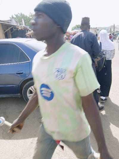 The image depicts an outdoor street scene in Mumbwa, Zambia, under bright daylight. In the foreground, a young Black male is captured in motion, facing right. He wears a black beanie, a light green and white striped short-sleeve shirt with a blue circular logo and another emblem on the chest, and light-colored jeans. A piece of red fabric is visible near his right hip. He holds a light-colored wooden stick with a white and blue cylindrical object at its end in his right hand. The subject exhibits motion blur, indicating movement.
To the left of the main subject, a dark blue sedan, possibly a Honda Accord, is parked or moving slowly. Further left in the background are rudimentary market stalls or buildings with light-colored walls and dark roofs.
In the mid-ground to the right, behind the main subject, an adult male walks away from the camera, wearing a dark garment and a patterned kufi-style cap. Behind him, an adult female also walks away from the camera, wearing a white headscarf, a dark top with white patterns, and dark trousers or a skirt, carrying a dark handbag on her right shoulder. Additional blurred figures are visible further in the background, suggesting a moderately crowded area. The ground surface is unpaved, appearing as dirt or gravel.