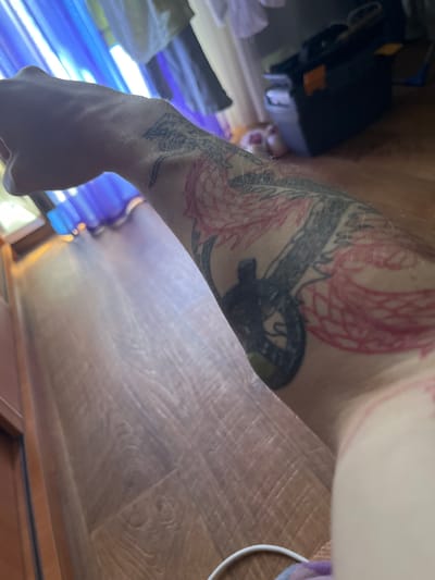 The image shows a close-up view of a person's tattooed arm. The tattoo appears to be a depiction of a dragon with pink and black ink. The arm is resting on a wooden floor. In the background, there are blue curtains and a dark toolbox. The setting is a residential interior. There are no discernible landmarks or context to suggest a specific location within Muravlenko, Russia.
