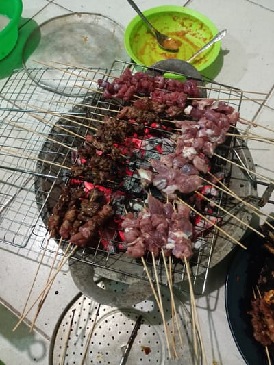 The image shows a grill with several satay skewers cooking over hot coals. Some skewers have raw meat, others are partially cooked, and some are well-done or slightly charred. The grill is placed on a stone or concrete surface, possibly a patio or outdoor area. A green bowl containing sauce is visible at the top of the image, along with a spoon. A metal tray and a plate with cooked satay are also present. The setting appears to be a home or small business, given the casual arrangement. The image was taken in Batam, Indonesia.