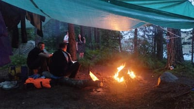 The image depicts a campsite in Tarasovskoe, Russia, with a group of people gathered around a bonfire under a makeshift shelter. The shelter is constructed from a large, teal tarp supported by poles and trees. In the foreground, two men are seated on the ground, facing the fire. One man, wearing a black jacket, is looking towards the camera. The other man, in a black shirt, is looking away from the camera. To the left of the men, there are several bags and a pair of shoes. Further back, a woman in a black and white patterned dress is standing, and another person in a white shirt with red trim is visible behind her. The environment is a forest with tall trees and dense foliage. The bonfire is burning brightly in the center of the scene, casting a warm glow on the surroundings. The ground is covered in dirt and leaves.
