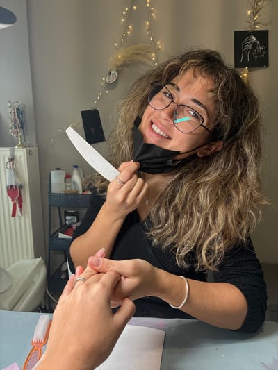 A woman with glasses smiles while holding a nail file and face mask. She wears a black top and a silver bracelet. Her long, wavy hair is brown with blonde highlights. She appears to be giving a manicure to another person, whose hands are visible, showing manicured nails. The scene takes place indoors with decorations including string lights and artwork on the wall behind her. A radiator and various bottles are also visible in the background.