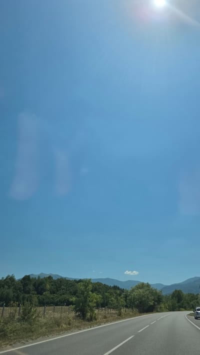 The image depicts a scenic view of a rural road in the vicinity of Mezdreya, Bulgaria. The road curves to the right and is bordered by a grassy shoulder and a fence. Beyond the fence, a field with dry grass and scattered trees leads to a dense forest. In the background, rolling hills and mountains are visible under a clear blue sky with the sun prominently shining in the upper right corner. The overall impression is one of a sunny, tranquil day in the Bulgarian countryside.
