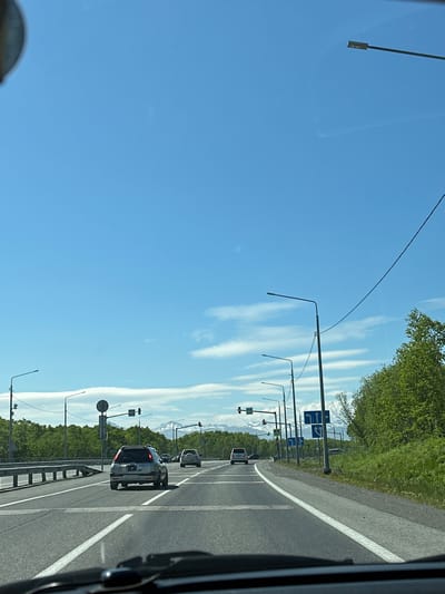 The image depicts a view from a vehicle traveling on a multi-lane road in the city of Svetly, Russia. The sky is clear blue with scattered white clouds. Several streetlights are visible along the roadside. On the right side of the road, lush green trees and vegetation line the embankment, with distant snow-capped mountains visible on the horizon. Traffic signs indicating lane directions are posted. Multiple vehicles are driving on the road, including a silver car in the foreground with a license plate that appears to read "B 557 PP 41". The road itself is asphalt with clear white lane markings. A metal guardrail is present on the left side of the road.