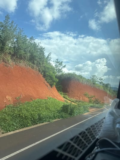 The image depicts a roadside view in An Phu Ward, Vietnam, with a focus on a red, eroded hillside adjacent to a paved road. Lush green vegetation, including bushes and trees, grows on top of and along the base of the hillside. The road has a white dividing line and yellow markings indicating a crosswalk or intersection. The sky is partly cloudy with bright blue patches. The scene is captured from the perspective of someone inside a vehicle, as evidenced by the reflection on the windshield and the presence of interior car elements, including a shoe, in the foreground.