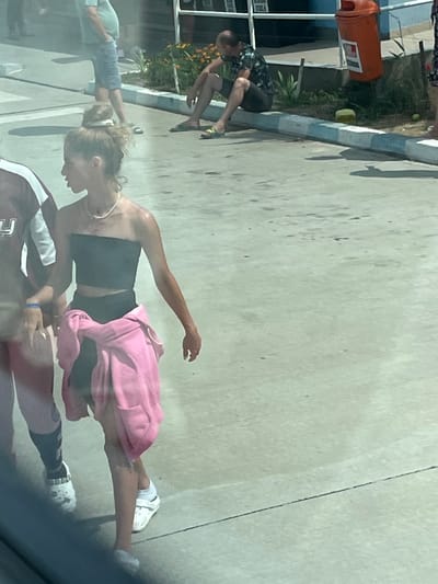 The image depicts an outdoor scene, likely in Ovidiu, Romania. In the foreground, a young woman with her hair pulled up walks towards the right. She is wearing a black strapless top, black leggings, and a pink hooded sweatshirt tied around her waist. She also wears white clogs and a necklace. To her left, partially visible, is another person wearing a maroon and white shirt and pink pants.
In the middle ground, a man is sitting on a concrete curb with his legs extended. He is wearing a dark floral print shirt and dark shorts, and appears to be looking down. He is wearing sandals. Behind him, to the right, is an orange trash can.
In the background, other individuals are visible, along with foliage and a building structure. The ground is paved, with some patches of dirt and grass visible. The overall lighting suggests it is daytime.