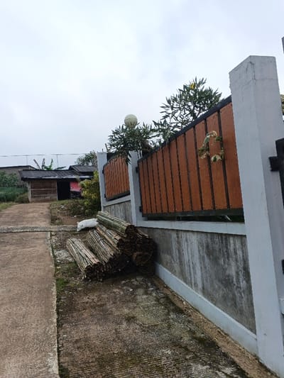 The image depicts an outdoor scene in Clacuc, Timor-Leste, under an overcast sky. In the foreground, a textured concrete path or driveway extends from the lower left, merging with an unpaved dirt area to the right. Leaning against a concrete wall on the right side of the frame is a stack of approximately ten bundles of cut bamboo or similar natural wood poles, secured with ties. A white plastic bag is visible on the ground near the base of the stack. The concrete wall, exhibiting weathering and discoloration, supports a modern fence section composed of dark brown vertical panels set within a black metal frame. Two light grey concrete pillars flank this fence section; the left pillar is topped with a spherical white outdoor lamp. Green foliage from bushes and trees is visible both behind and above the fence structure. In the distant background to the left, a single-story building with a dark-pitched roof and an open doorway is visible. A faint red object is partially discernible behind this building. No individuals are present in the scene.
