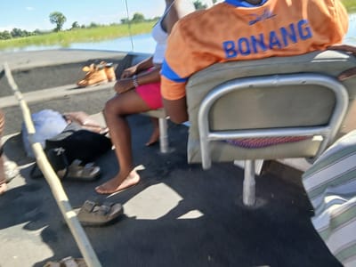 The image shows a group of people on a boat. One person, wearing an orange shirt with the word "BONANG" printed on it, is sitting on a metal-framed chair. Another person has bare feet and is wearing pink shorts. There are several pairs of sandals scattered around the boat floor. A long wooden pole lies across the boat. The background reveals a grassy shoreline with scattered trees under a blue sky, suggestive of Gunotsoga, Botswana.