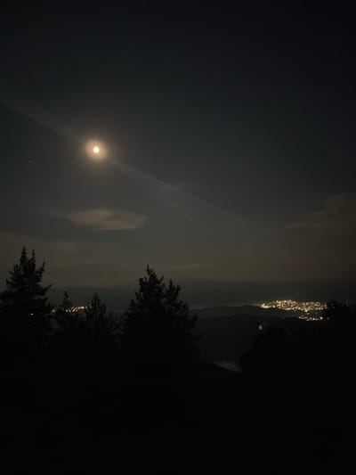 This is a night photograph, taken in Simeonovets, Bulgaria, under a dark, cloudy sky. The full moon is visible in the left portion of the frame, casting a soft glow that illuminates some of the clouds. A faint beam of light extends from the moon, creating a subtle lens flare effect. In the distance, on the right side of the image, there are faint lights that appear to be from a settlement or buildings, possibly on a hillside. Silhouetted trees are visible in the foreground on the right, with their dark foliage contrasting against the faintly lit sky. The overall mood of the image is serene and tranquil, suggesting a quiet night in a rural setting. No people or specific objects are clearly identifiable, beyond the natural elements of the moon, clouds, and trees, and the distant artificial lights. The time of day is clearly night, indicated by the darkness and the presence of the moon and stars. The cloudy conditions suggest potentially damp or humid weather.