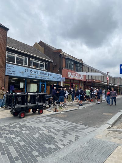 The image depicts an outdoor scene on a street in Broxbourne, United Kingdom, likely in a commercial area with shops. The sky is overcast. In the foreground, to the left, a dolly with stacked metal equipment is positioned on a patterned paved area. To the right, a group of people, appearing to be a film crew, are gathered around various pieces of equipment, including cameras, microphones, and lighting stands, on a section of asphalt road.
Several shops are visible in the background. The most prominent is "ST JOHN'S CHARITY SHOP," identifiable by a large blue sign above its storefront. Next to it, to the right, is a shop labeled "BAZAD VIDEO HOME VIDEO RENTAL." Further down the street, other shop signs are partially visible, including "JACK'S CAFE," "THE FERTILITY FOUNDATION," and "RONA." People are standing on the pavement in front of these shops, some interacting with the film crew. The overall atmosphere suggests a film or television production is underway on a public street.