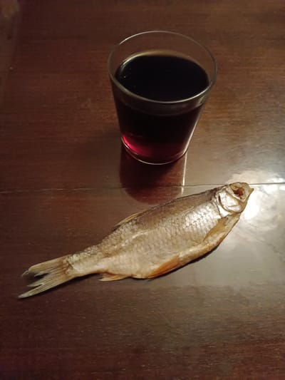 A still-life composition on a dark wooden surface features a clear glass tumbler filled with a dark liquid, possibly red wine or cola. The liquid reaches near the top of the glass. In the foreground, a dried, pale-colored fish rests next to the glass. The fish's features, including the head, scales, and fins, are clearly visible. The setting appears to be indoors. There is no indication or suggestion that this image was taken in Verkhnii Dvor, Russia.