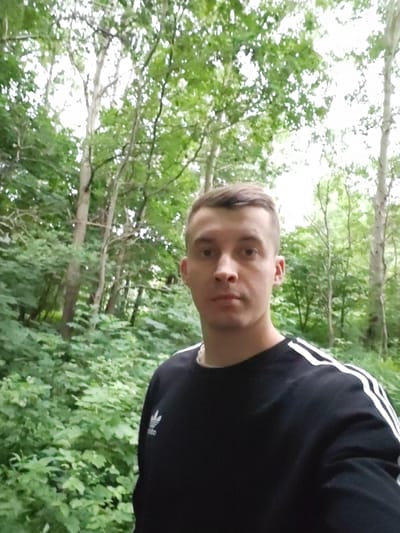 A young man is captured in a selfie-style shot, positioned in the foreground against a backdrop of dense green foliage and trees, suggesting an outdoor, possibly wooded or park-like setting. He is wearing a black sweatshirt with white stripes on the shoulders and a silver necklace. His facial expression is neutral, and he is looking directly at the camera. The lighting indicates it is daytime, with diffused natural light filtering through the leaves. The specific location within the city of Васильково, Russia, cannot be determined from the visual information alone.