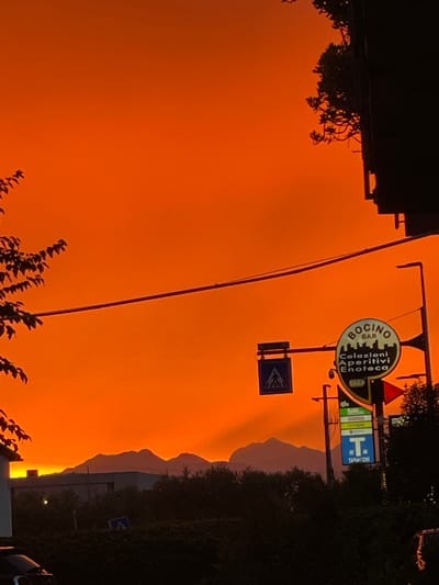 This media file captures a striking sunset scene in Castelfranco di Sotto, Italy. The sky is dominated by a vibrant, fiery orange hue, suggesting a dramatic atmospheric event, possibly due to smoke or dust. Silhouetted against this intense sky are the outlines of buildings on the left and a dense area of trees and foliage on the right.
Prominently displayed in the center-right of the image are several signs. The largest, circular sign features the text "BOCINO BAR Colazioni Aperitivi Enoteca" with an illustration of buildings. Below this, a blue square sign with a white pedestrian crossing symbol is visible. Further to the right, another blue sign displays a white "T" and the text "TABACCHI," indicating a tobacco shop.
The image is oriented vertically, with the top of the frame showing the dark silhouette of a building and what appears to be hanging branches or vines. The lower portion of the image reveals more of the dark foliage and a hint of a bright light source on the far right, likely from a streetlamp or building light. The silhouette of mountains or hills can be seen in the distance, their shapes softened by the hazy, orange atmosphere. The overall impression is one of an intense and possibly unusual sunset, casting a dramatic light over the urban and natural elements of the scene. The time of day is clearly evening, during sunset.