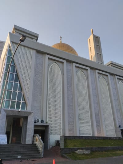 The image depicts a mosque in Sidodadi, Indonesia, characterized by its modern architecture. The building features a prominent golden dome at its peak, topped with a crescent moon symbol, and a tall, slender minaret with decorative openings. The facade is predominantly light gray, with textured panels featuring a repeating geometric pattern. Large arched windows with intricate latticework adorn the main structure.
In the foreground, a wide set of stone stairs leads up to the mosque entrance. To the left of the stairs, a sign with text is partially visible, along with a portion of a street lamp. The ground level appears to be paved with asphalt and stone, with a strip of green grass and a low stone wall separating the paved area from the building. Two outdoor air conditioning units are visible on the side of the building near an entrance. The sky is clear and pale blue, indicating daylight. No people are clearly visible in the image, though there are some shoes on the ground near the stairs.