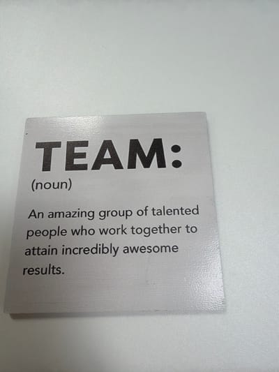 A close-up image shows a card lying on a white surface. The card has the word "TEAM:" printed in large, bold, black letters. Below "TEAM:" is "(noun)" in a smaller font. To the right of "TEAM:" is a definition that reads: "An amazing group of talented people who work together to attain incredibly awesome results." The card is white with a slightly textured surface. The image is not focused in a place. It is difficult to ascertain the exact location without further information about the venue. No individuals or interactions are visible in the image.