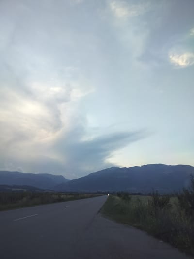 The image depicts a rural scene in Manolovo, Bulgaria, at dusk. A paved road stretches into the distance, flanked by grassy fields and low-lying vegetation. In the background, a range of dark, silhouetted mountains forms the horizon. The sky is vast and cloudy, with dramatic formations of white and grey clouds that create a sense of depth and atmosphere. The overall mood is serene and somewhat dramatic, possibly due to the approaching or receding storm suggested by the cloud formations. The lighting indicates that it is either early morning or late evening, with the sun likely low on the horizon. There are no people or specific objects visible besides the road and natural landscape. No text is present in the image.