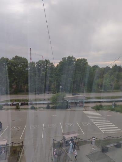 The image shows a view from above, looking out over a road and a parking lot. In the distance, two tall industrial smokestacks with red and white bands emit light smoke into an overcast sky. A line of dense green trees borders the road.
In the foreground, a paved parking lot has several marked parking spaces, four of which are designated for disabled individuals, indicated by the International Symbol of Access. A pedestrian crosswalk is clearly marked, leading across the parking lot towards a bus stop shelter. Several people are visible in the scene: on the swings under a canopy, standing near the bus stop, and walking in the parking lot. A bench is situated in the lower right corner of the image. A parked car is partially visible on the left side of the parking lot.
The scene appears to be located near an industrial area, suggested by the smokestacks, and a public space with a road, parking, and public transport facilities.
