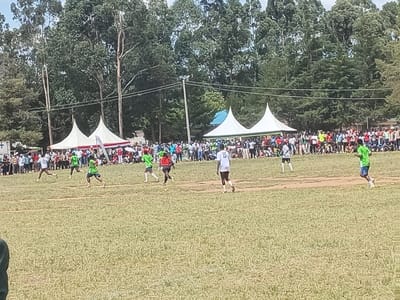 The scene depicts an outdoor football match in progress at Manda-Shivanga ward, Kenya. Multiple players are visible on a predominantly grassy field with some bare dirt patches. Players on the left are primarily wearing green jerseys with black shorts, actively running and positioned in mid-action. One player in a red jersey and black shorts is also visible. On the right, players are wearing white jerseys with dark shorts and green jerseys with black shorts, also in motion. A large crowd of spectators lines the far side of the field, standing and seated, observing the game. In the background, behind the spectators, are several white conical tents and at least one blue tent. A dense line of tall green trees forms the horizon, with power lines and poles visible against them. A white sign with a red cross is partially visible on the far left. The weather appears bright and clear.