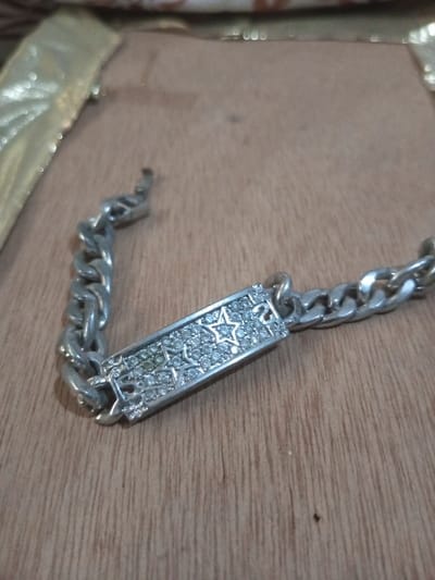 A silver-colored chain bracelet with a rectangular ID plate is depicted, resting on a light brown wooden surface. The ID plate is encrusted with numerous small, clear, faceted stones, featuring two five-pointed star outlines within the pavé setting. The chain is a curb link style, and the bracelet appears unclasped, with the clasp end visible on the left. In the blurred background, a segment of a gold-colored, reflective object is partially visible on the upper left. This scene is situated in the city of Chandragiri, Nepal.