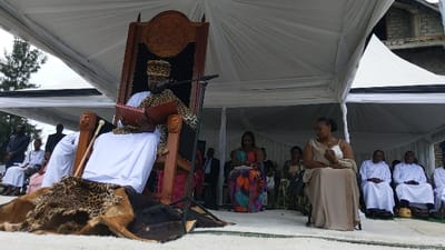 The image depicts a man, possibly a king, in a white robe and a leopard-print vest, sitting on a carved wooden throne under a white canopy tent. He is reading from a book, and a microphone is attached to the throne. He is seated on a leopard and cowhide rug. Other attendees are seated in rows behind him, some wearing traditional clothing. The scene appears to be an outdoor ceremony, possibly a coronation or a royal address. The location is Rukoko, Democratic Republic of the Congo, with the coordinates: -1.624, 29.258. The environment is a grassy field under white canopies, and there is a stone building in the background.