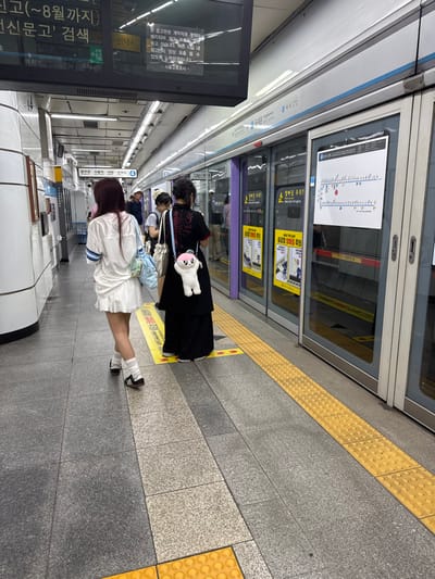 The image depicts an underground subway platform at Jongno 3-ga Station in Jongno-gu, Seoul, South Korea. Two individuals are prominently featured from behind on the grey-tiled platform. The individual on the left wears a white short-sleeved top with blue trim, a white pleated mini-skirt, white ankle socks, and black sneakers, walking towards the right side of the frame. This person carries a light blue shoulder bag. The individual on the right stands closer to the platform edge, facing a series of purple-tinted glass platform screen doors. This person wears a long black garment and carries a light blue shoulder bag from which a white, plush, rounded character with pink cheeks hangs.
The platform floor includes a continuous yellow tactile paving strip running parallel to the screen doors. Yellow directional arrows are present on this strip. The platform screen doors are adorned with multiple yellow warning signs displaying Korean text, notably "발빠짐 주의!" (Mind the gap!), and a white poster illustrating a subway line map. Overhead, a large black digital display screen shows Korean text, including "종로3가" (Jongno 3-ga) and "동대문" (Dongdaemun), alongside a green emergency exit sign. The station is brightly illuminated by overhead lighting fixtures. Several other blurred individuals are visible in the background along the platform.