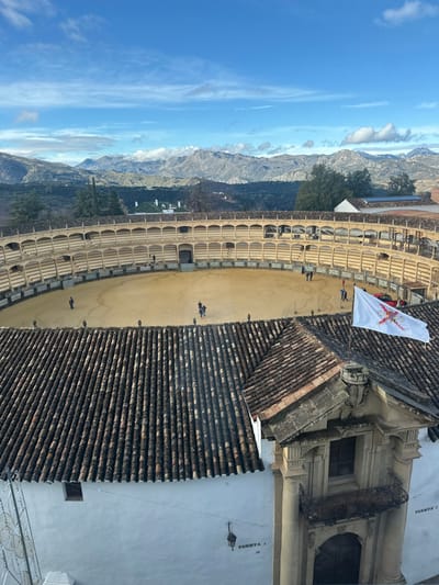 An elevated, wide-angle image captures the Plaza de Toros de Ronda in Ronda, Spain, under a partly cloudy blue sky. The central feature is the circular bullring, characterized by its light-brown, sandy arena floor. The surrounding spectator area consists of multi-tiered, arcaded stands constructed from light-colored stone or stucco, showing two distinct levels of arches. Approximately 10-15 individuals are observed in small groups on the arena floor, walking and standing.

In the foreground, the steeply sloped, terracotta-tiled roof of an adjacent building occupies the lower portion of the frame. An ornate, light-stone architectural structure with columns and a small, wrought-iron balcony is integrated into the building on the right, from which a white flag bearing a red Cross of Burgundy is flying. The white-painted wall of the building below displays the text "PUERTA 3" (Gate 3) in two locations and features a dark metal lantern hanging near one inscription.

Beyond the bullring, rugged, green-grey mountains dominate the distant horizon, partially veiled by wispy white clouds. Verdant trees and scattered white buildings with red-tiled roofs are visible between the bullring structure and the mountain range. The scene is illuminated by natural daylight.